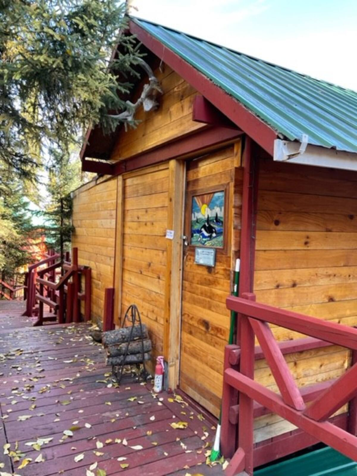Wooden cabin with green metal roof and red railings on a leaf-covered deck in the woods