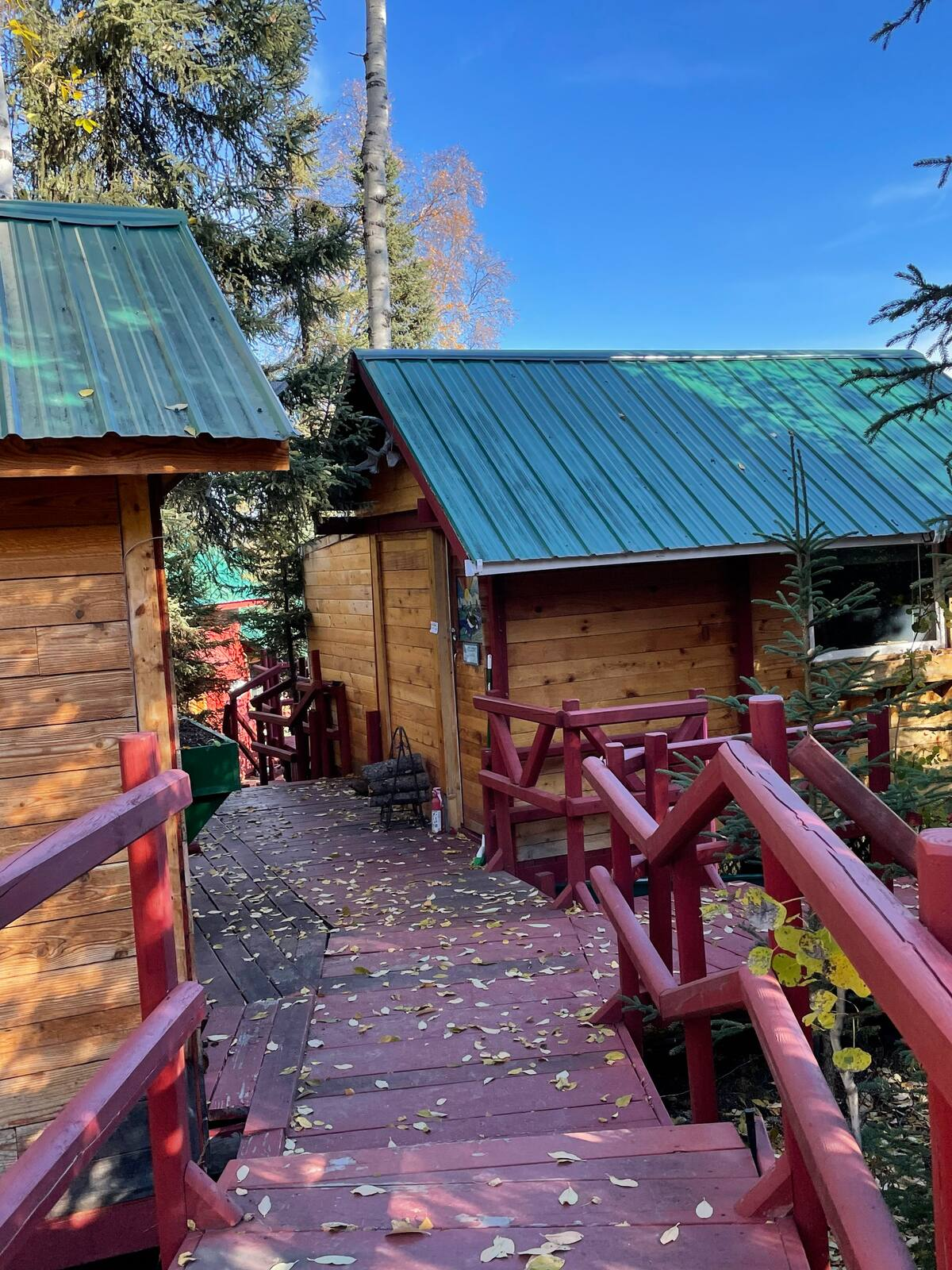 Wooden cabin walkway with red railings, green metal roofs, and trees under a blue sky.