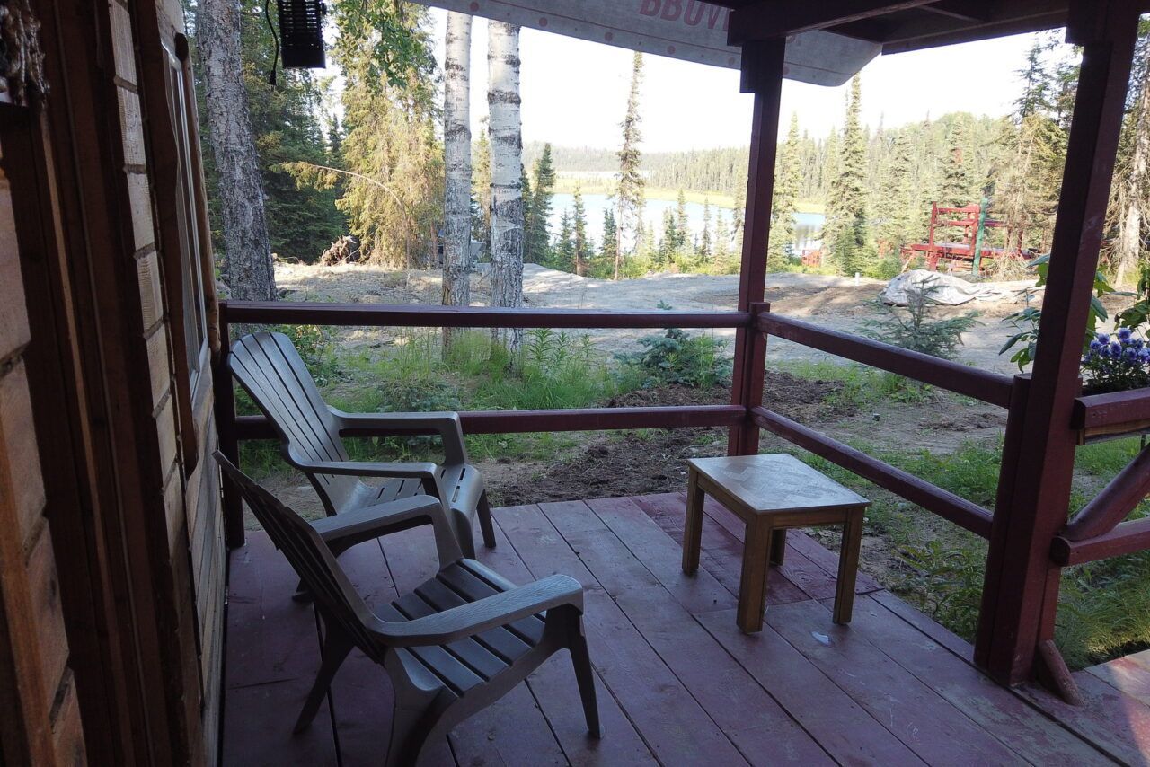 Covered porch with two chairs and a small table, overlooking a wooded yard and garden.