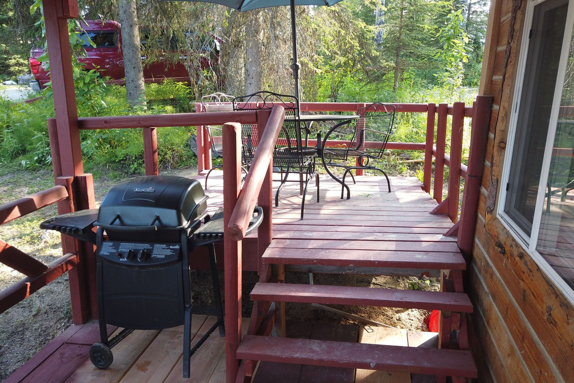 Wooden deck with a black grill, red railing, and patio chairs overlooking a wooded yard