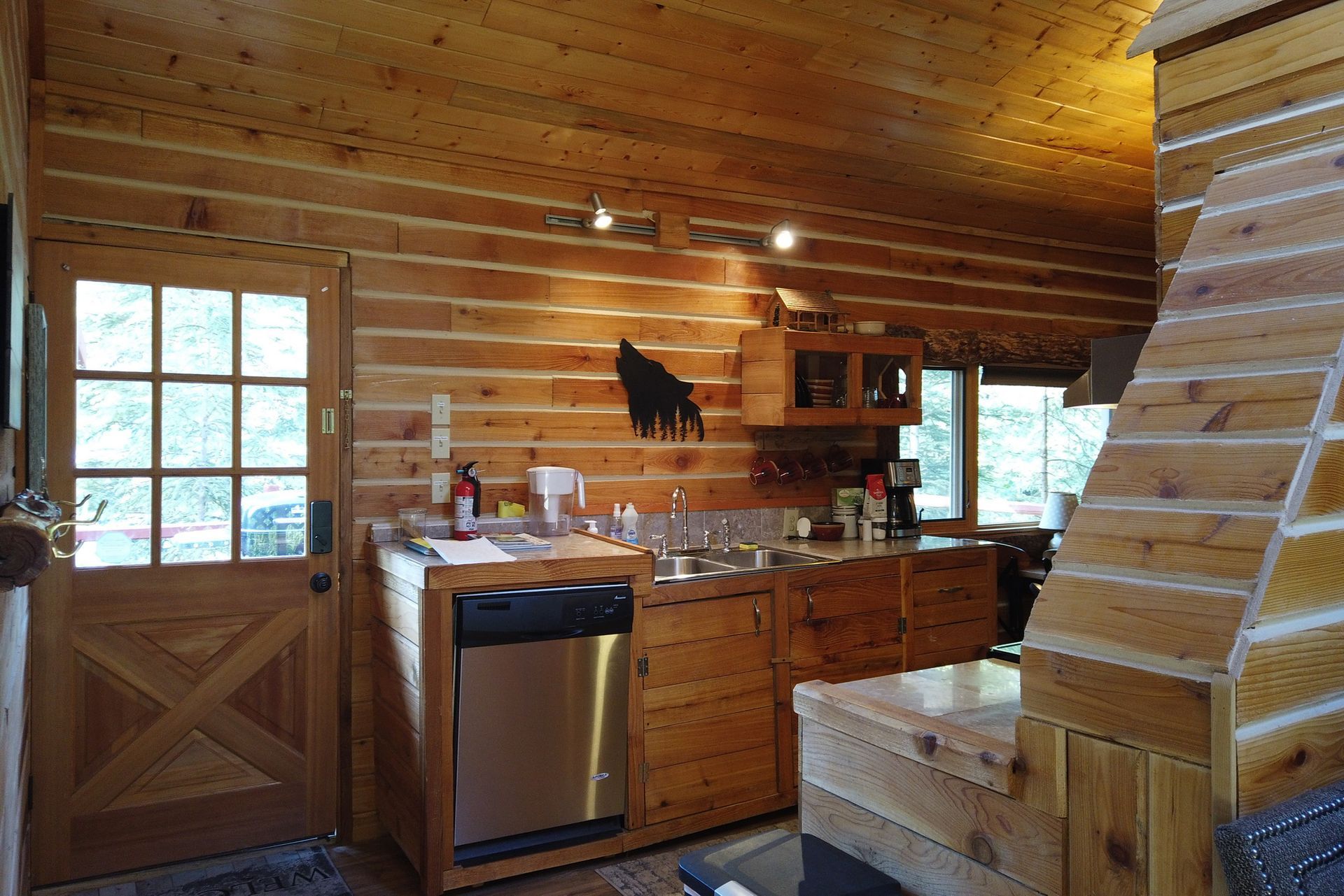 Rustic log cabin kitchen with wooden cabinets, stainless dishwasher, and loft stairs under a wood ceiling