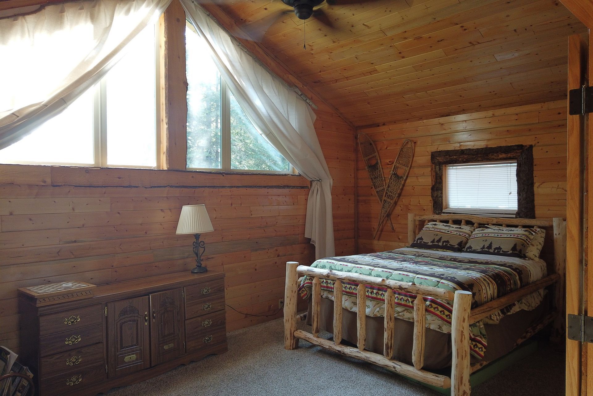 Rustic bedroom with wood-paneled walls, a wooden bed, dresser, and curtained windows under a vaulted ceiling