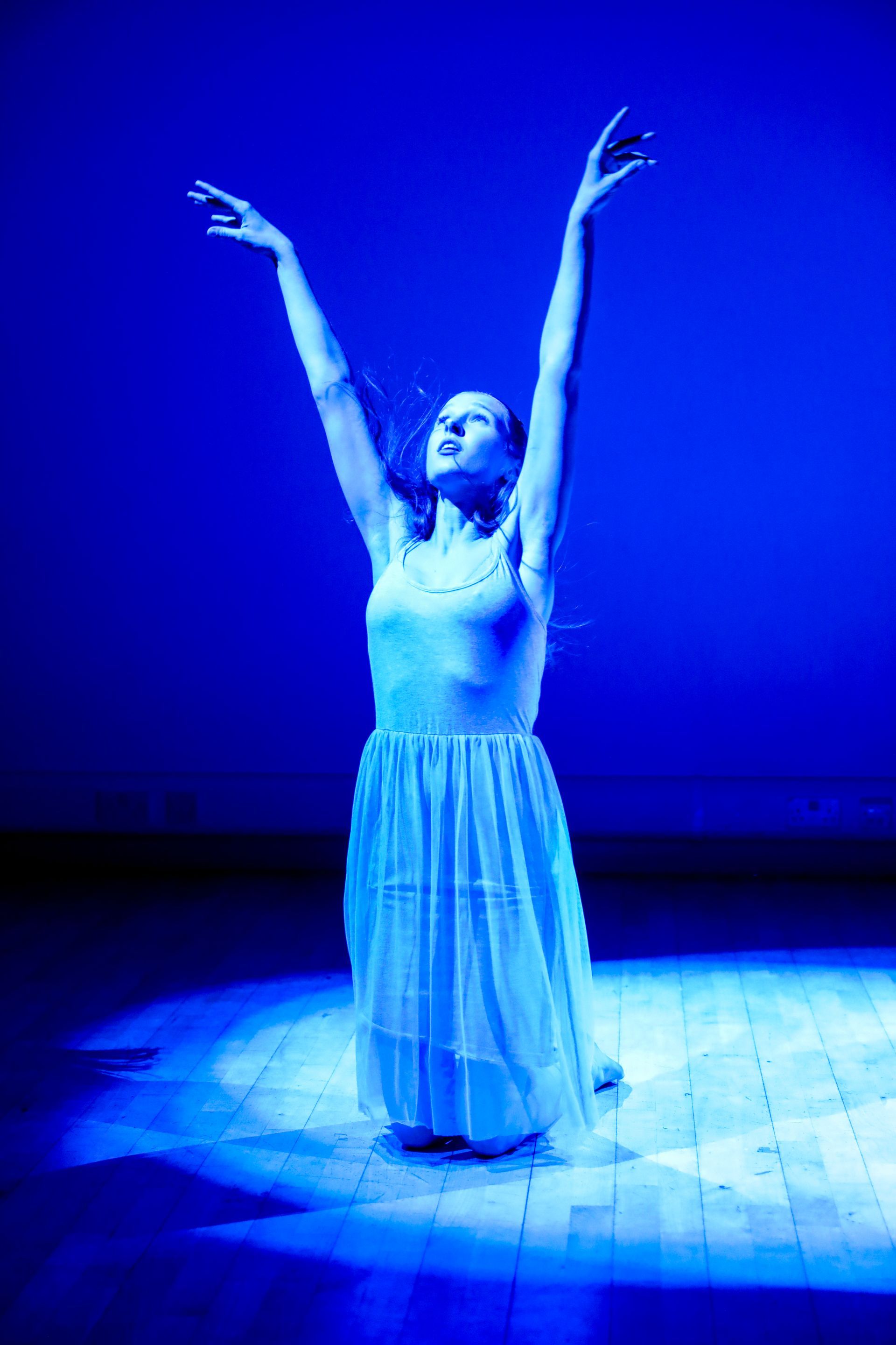 A woman in a blue dress is dancing on a stage with her arms in the air.
