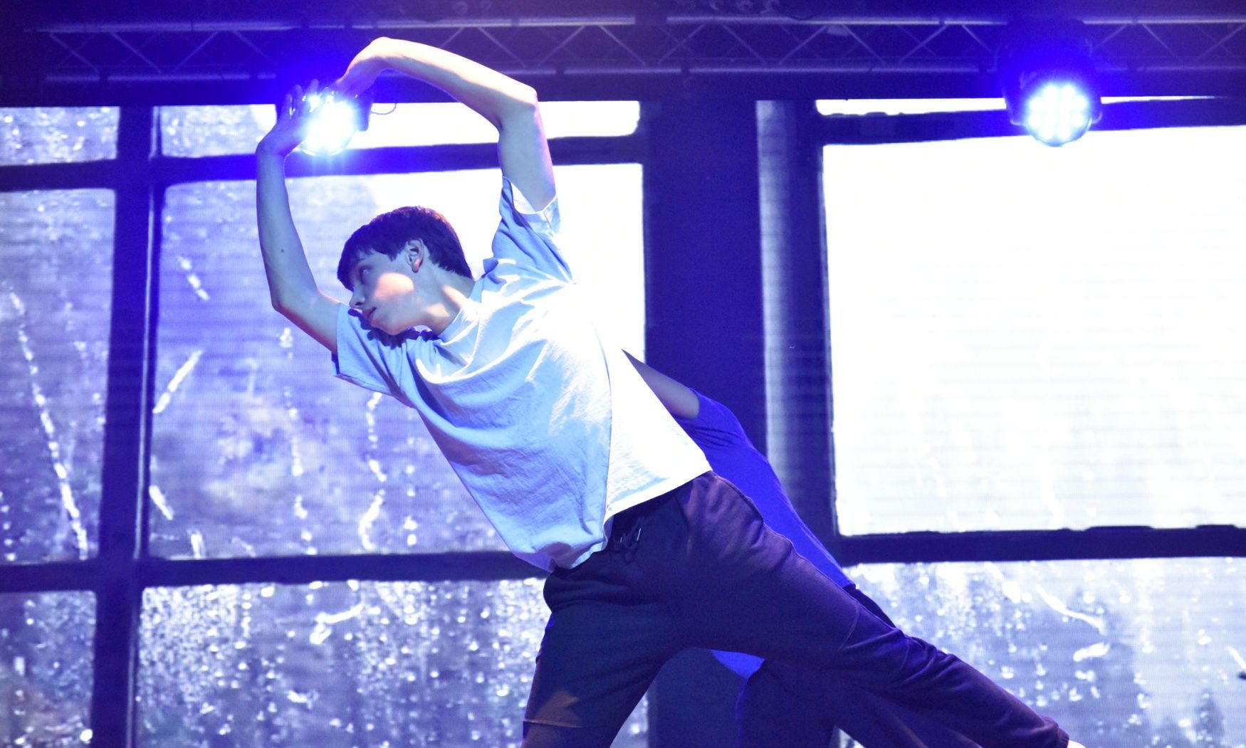 A man in a white shirt is dancing on a stage with a blue light behind him.