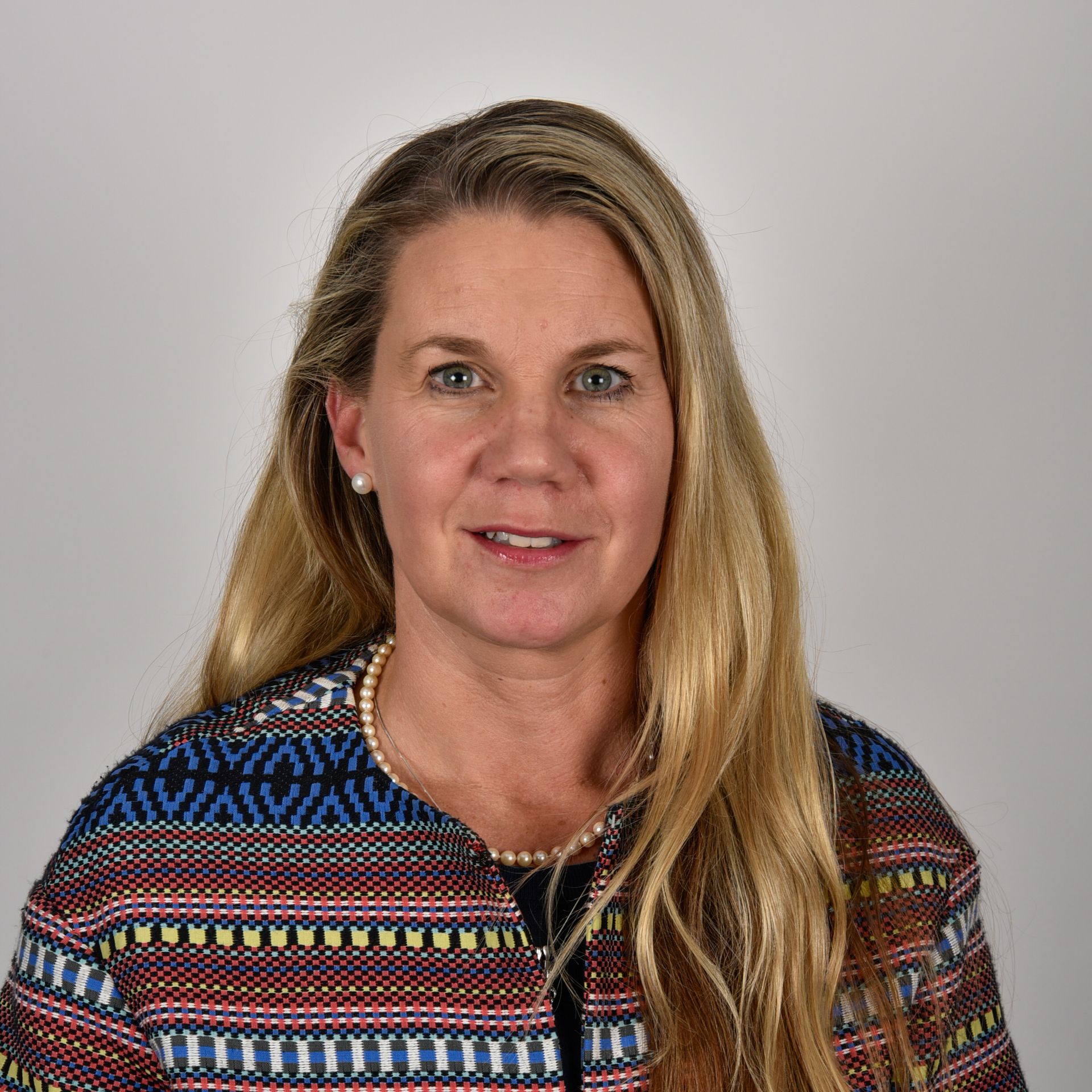 A woman with long blonde hair is wearing a colorful jacket and pearls.