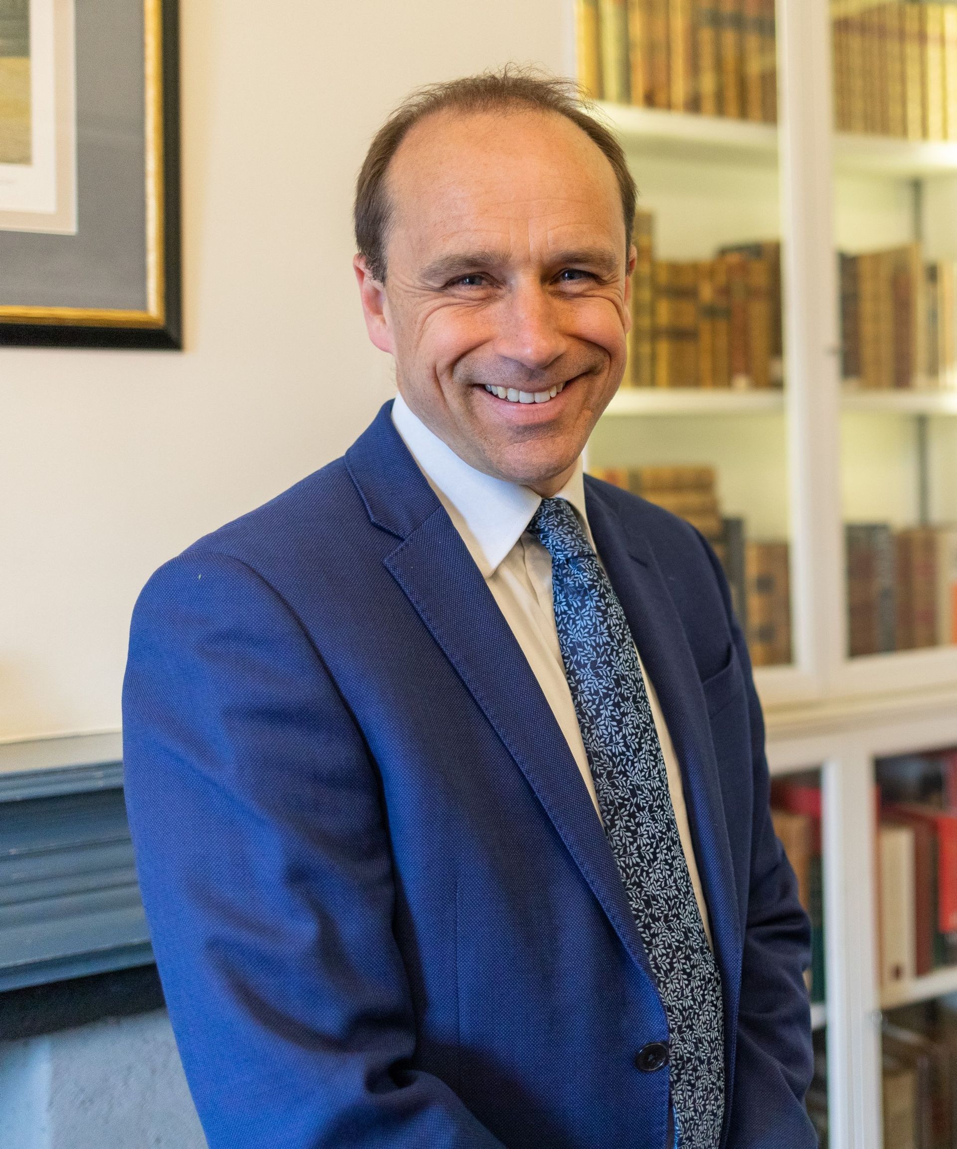 A man in a blue suit and tie is smiling in front of a bookshelf.