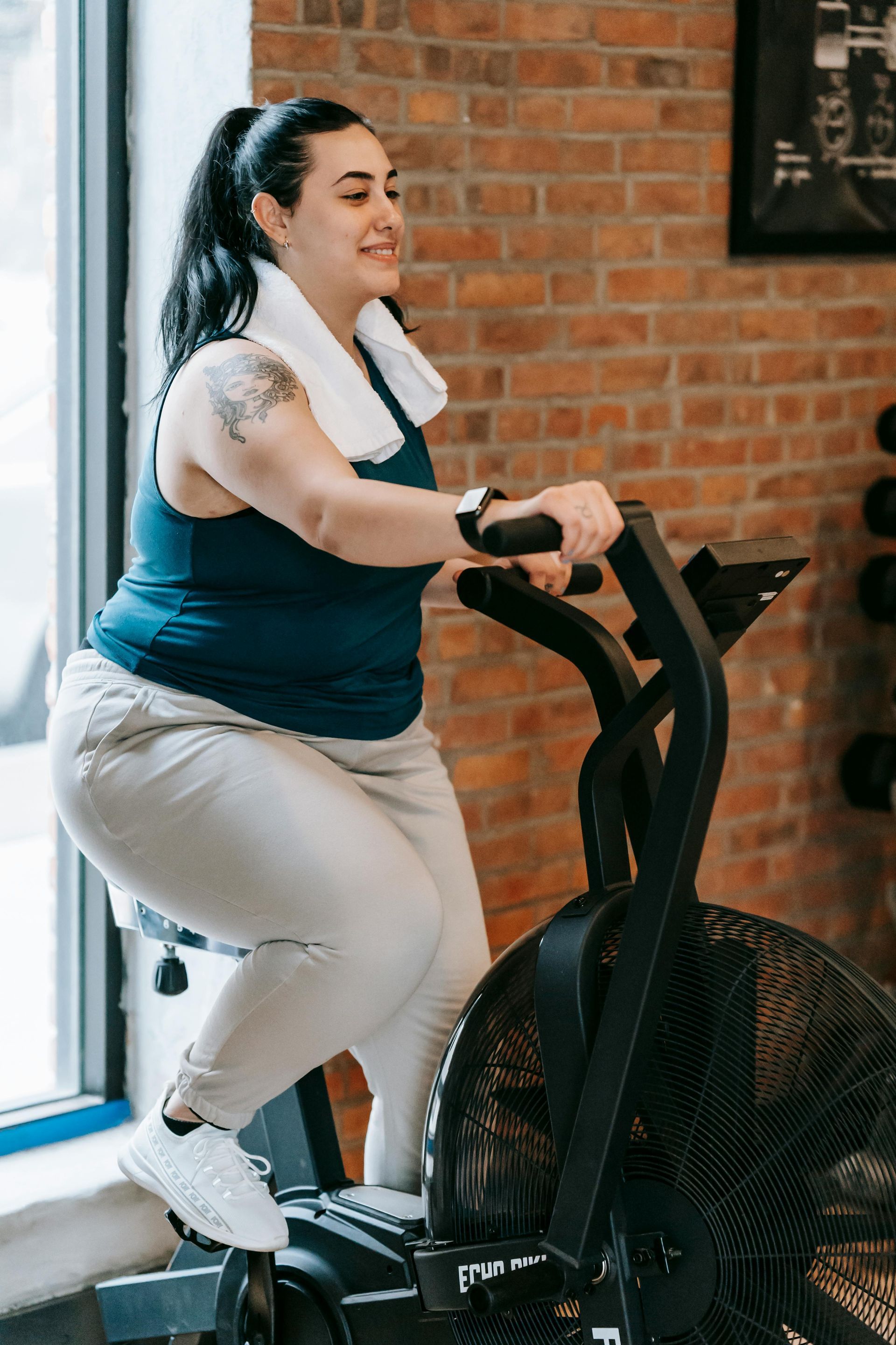 Woman exercising on an air bike, smiling, with a towel on her shoulders, in a gym.