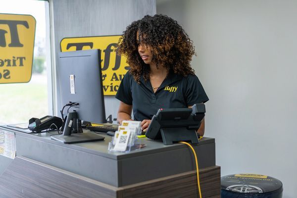 Woman at a desk in an auto shop, looking at a computer, with a cash register and equipment visible.
