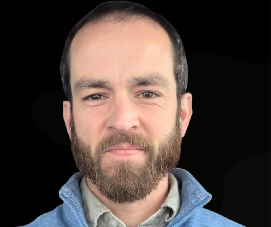 Man with brown beard and fair skin smiles, wearing a blue jacket and gray shirt, against black background.