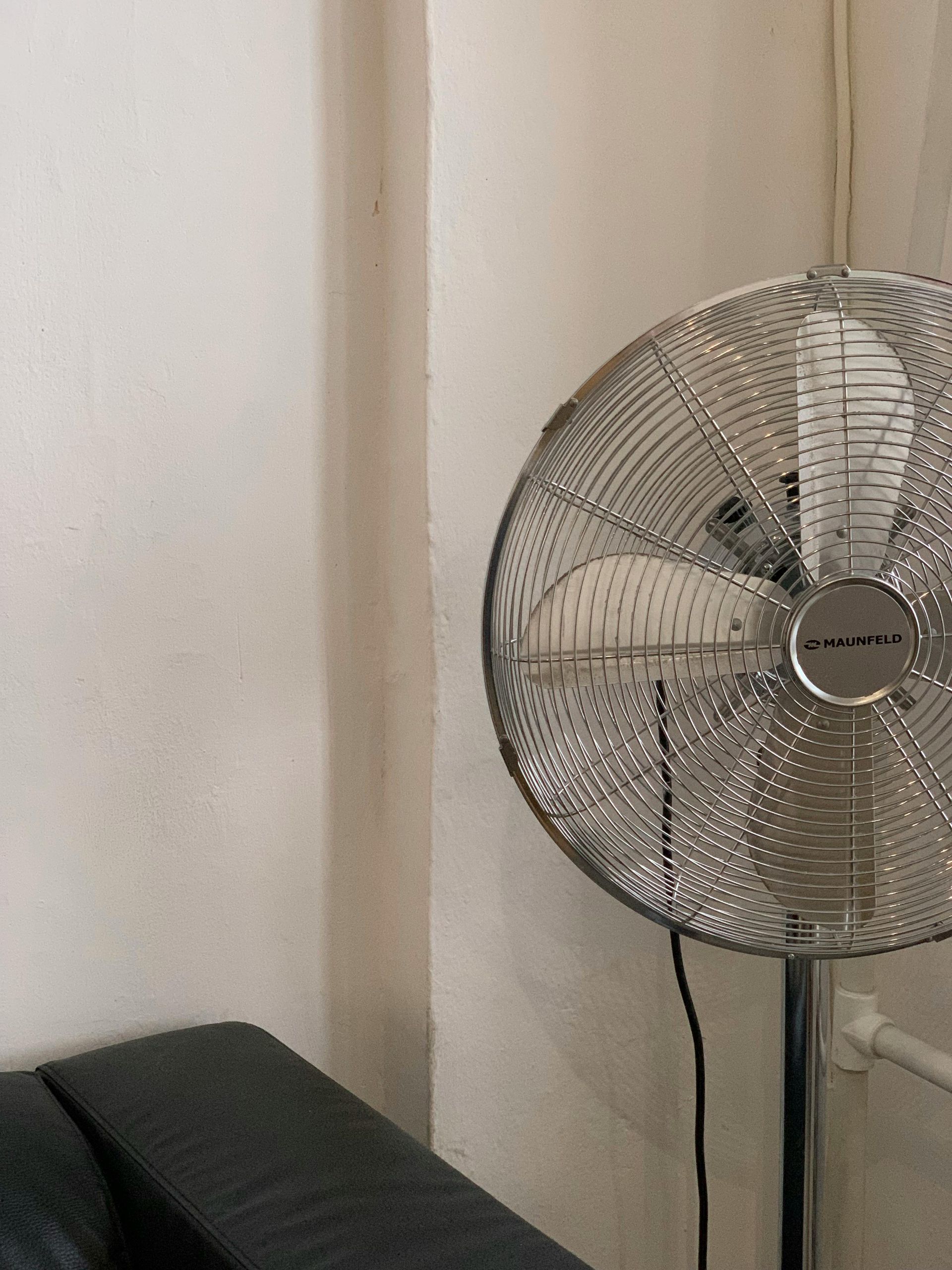 Silver oscillating fan in front of a white wall.