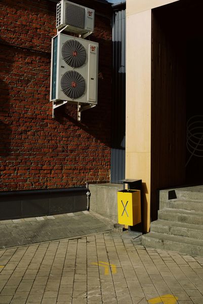 Yellow trash bin next to tan doorway, brick wall with air conditioner, brick paving.