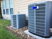 Two gray air conditioning units outside a beige house on a bed of rocks.