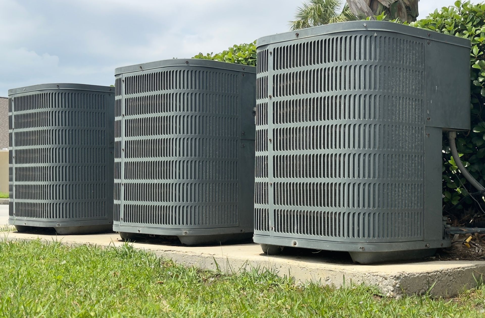 Three gray air conditioning units in a row on a concrete base, set in a grassy yard.