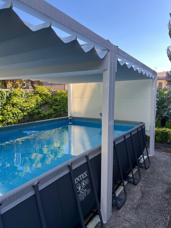 Piscina coperta nel cortile sul retro, con acqua azzurra, pali bianchi per la tettoia e lettini pieghevoli a bordo piscina.