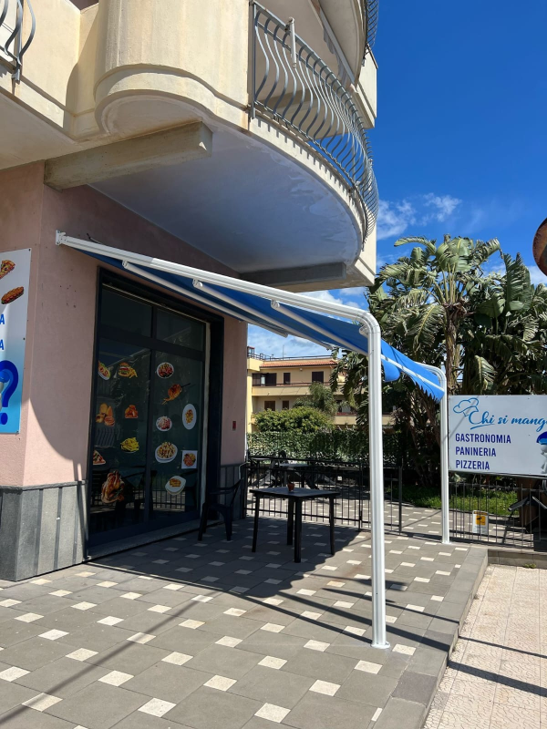 L'ingresso del negozio si trova sotto un balcone con una tenda da sole blu e un marciapiede piastrellato, accanto a un'insegna bianca.