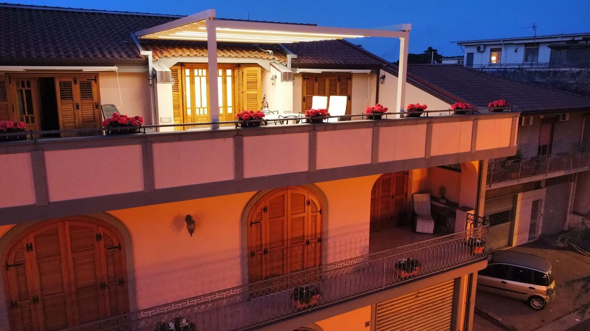 Vista al tramonto di una casa rosa a due piani con terrazza sul tetto e luci scintillanti, con un'auto parcheggiata al di sotto.