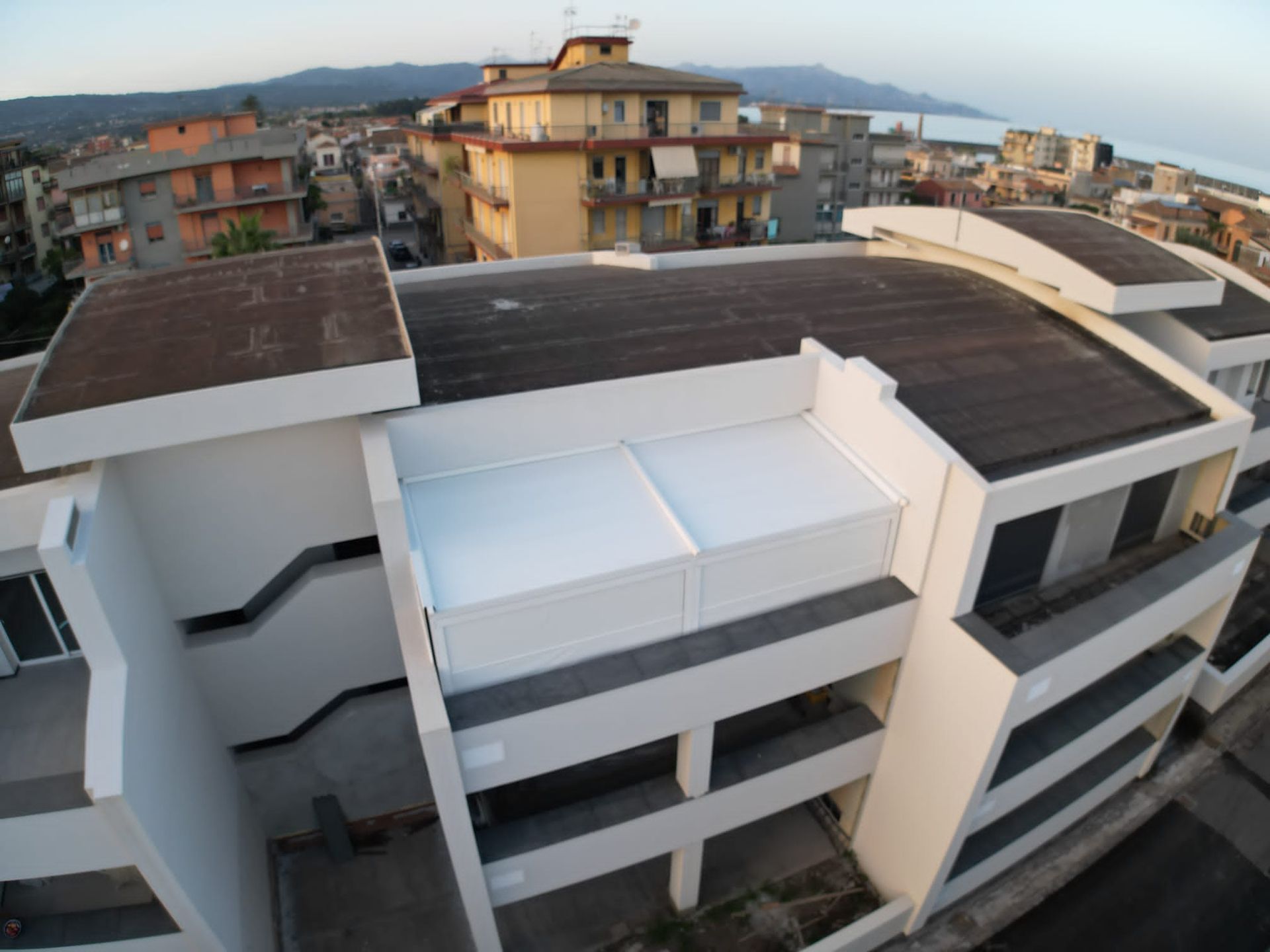 Veduta aerea dei moderni tetti bianchi di un palazzo che si affaccia su una cittadina costiera e sul mare al tramonto.