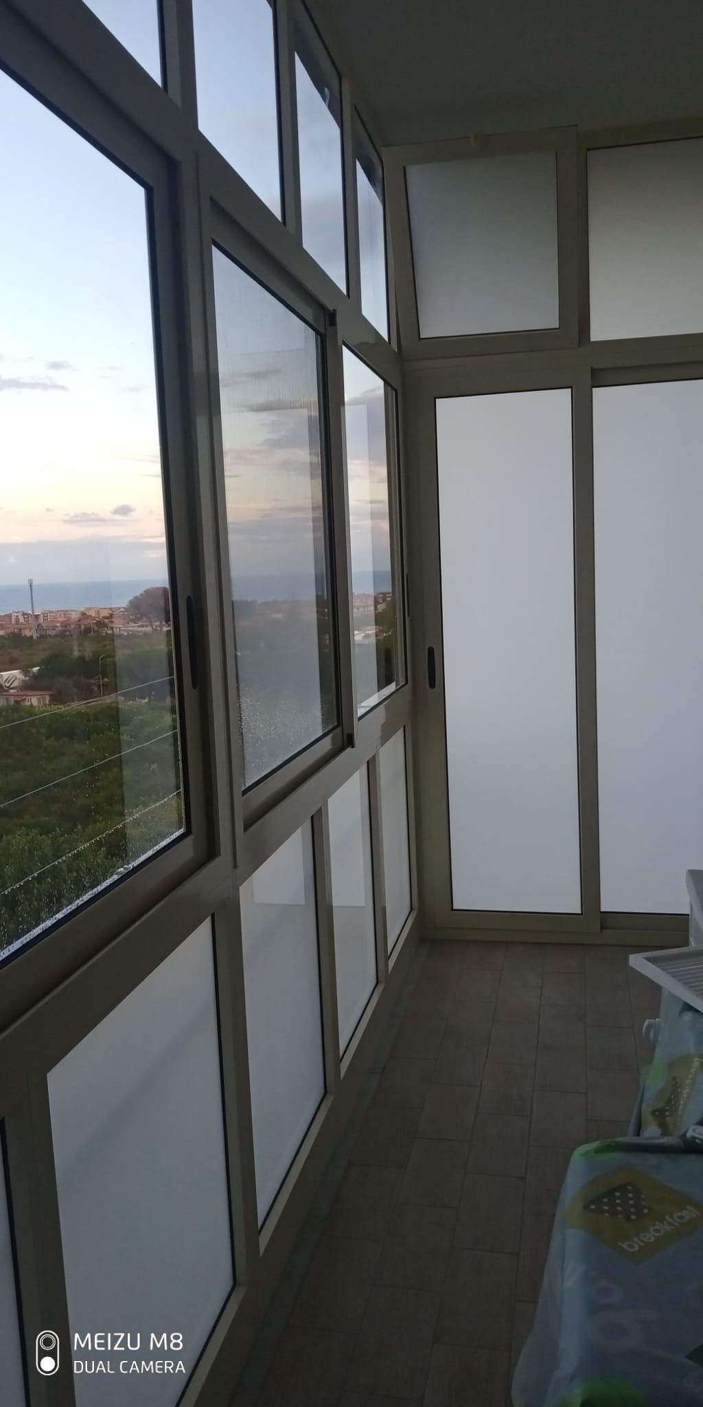Balcone vetrato con vista sulla città al tramonto e sedia appoggiata al muro.