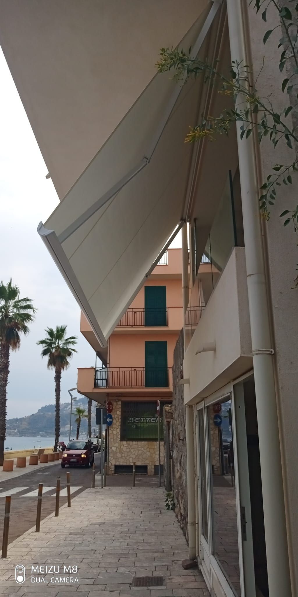 Marciapiede coperto accanto a un edificio moderno e palme, con auto parcheggiate lungo una strada sul lungomare.