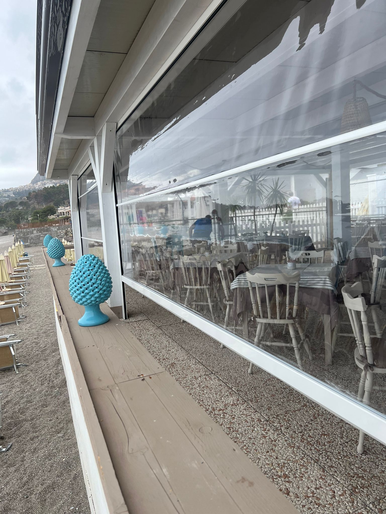 Esterno di un ristorante sul mare con ampie vetrate, tavoli all'aperto e una scultura decorativa blu lungo il percorso pedonale.