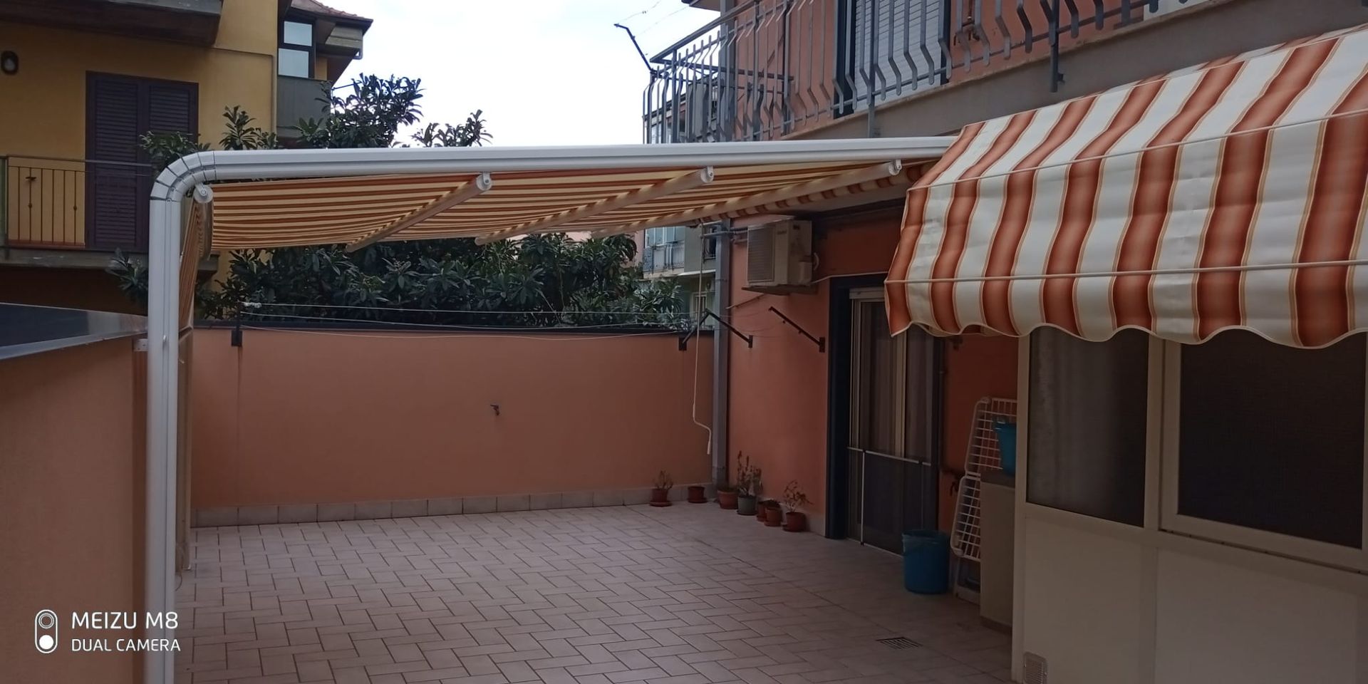Cortile con pavimento piastrellato, pergola in metallo e tenda a righe accanto a muri color pesca