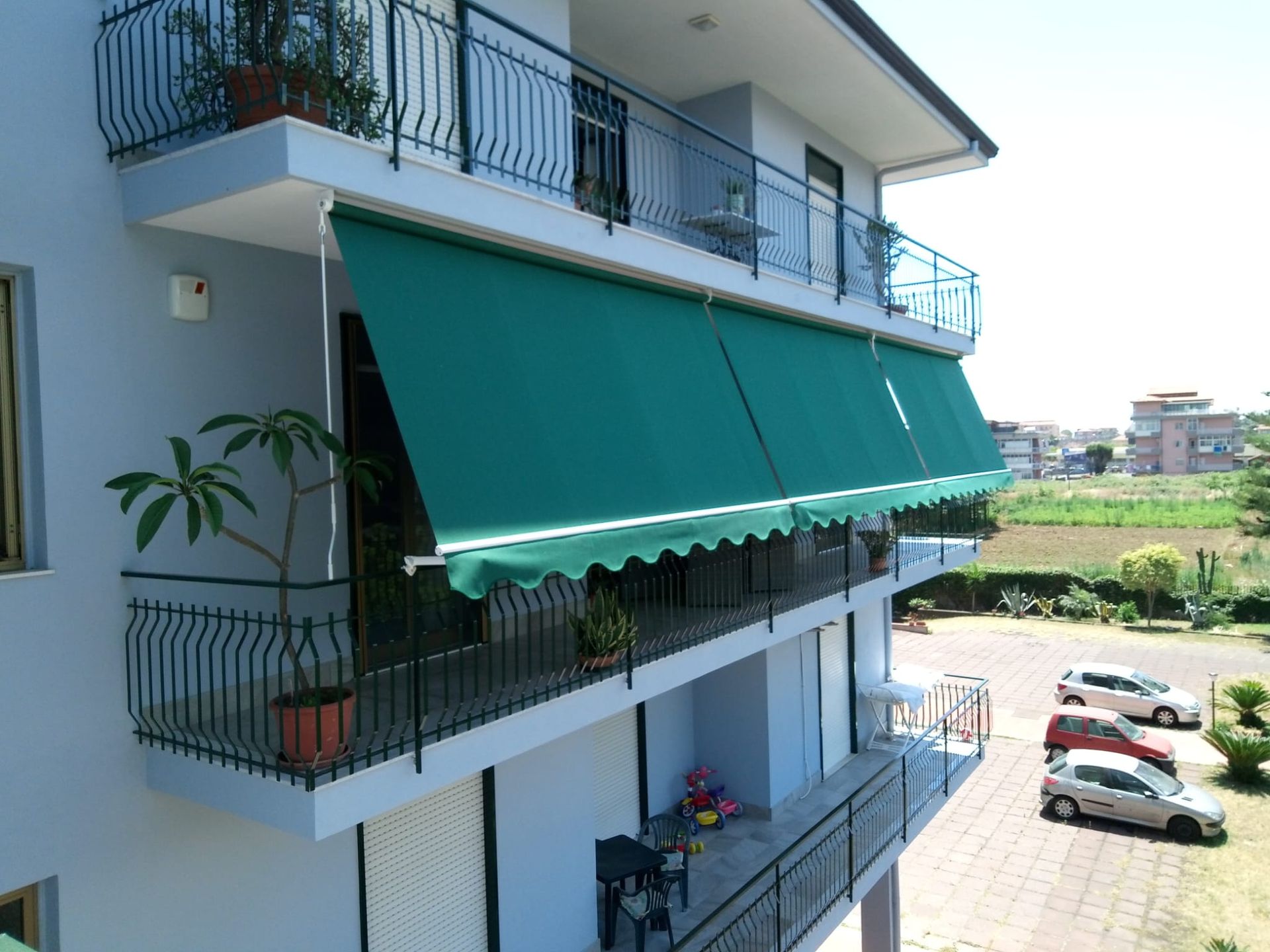 Palazzo residenziale con tende da sole verdi, balconi e auto parcheggiate in una strada soleggiata.