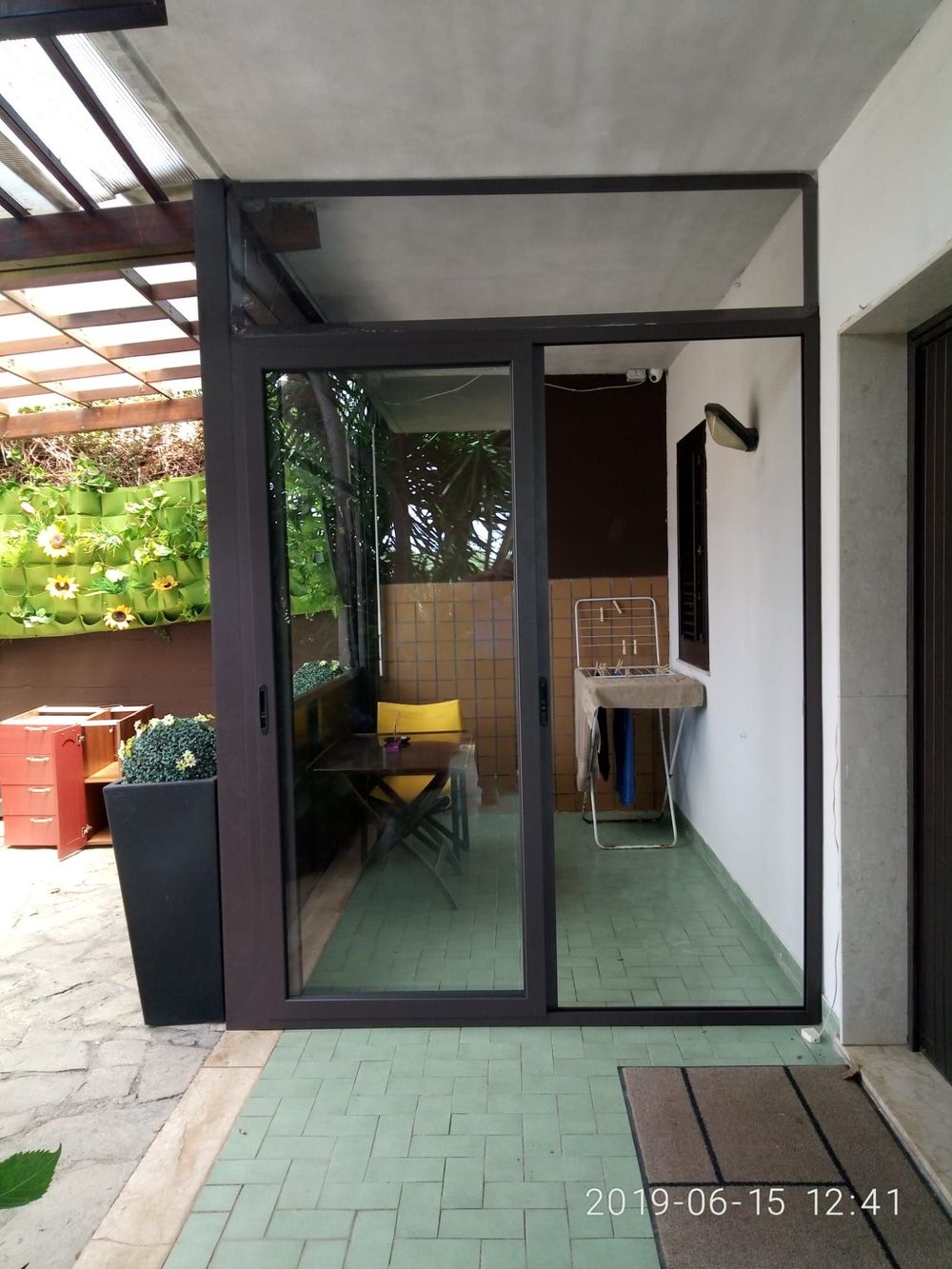 Ingresso con facciata in vetro e telaio nero, pavimento piastrellato verde e mobili da giardino visibili all'interno.