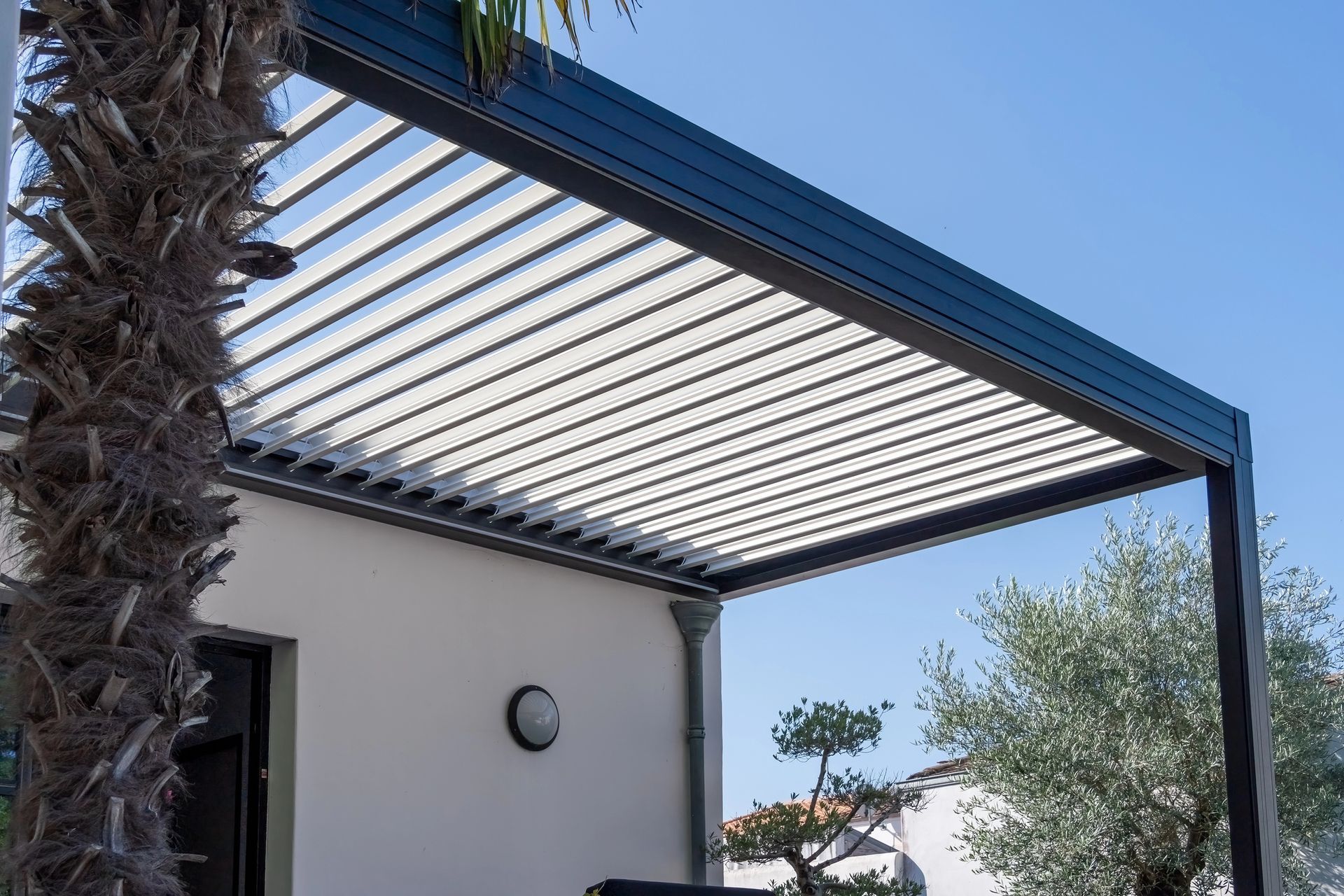 Patio con tettoia a pergola a doghe accanto a una casa bianca e una palma sotto un cielo azzurro.