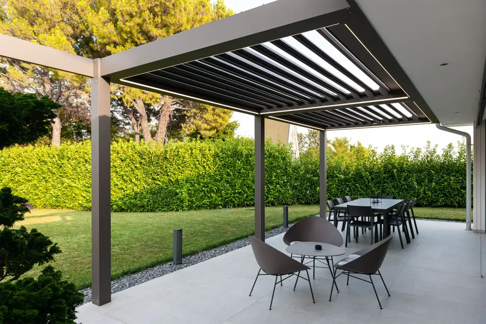 Patio coperto con tavolo da pranzo, sedie e piante in vaso, adiacente a una casa moderna e a un prato.