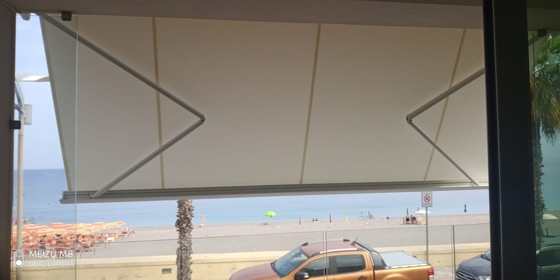 Vista sulla spiaggia attraverso una finestra con una tenda da sole grigia, mare blu e un'auto parcheggiata sottostante.