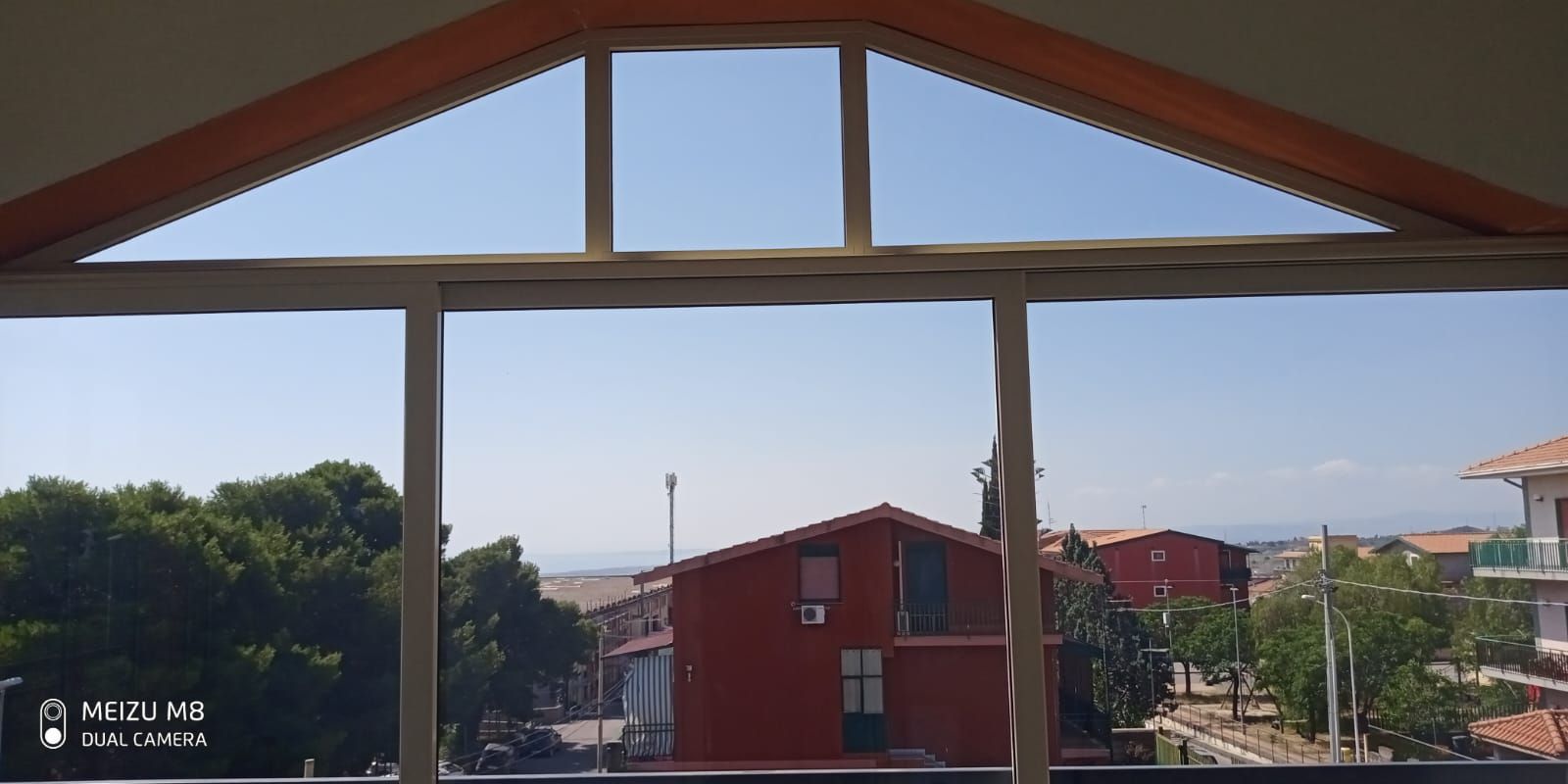 Edifici dai tetti rossi visti attraverso una finestra con cornice triangolare in legno, sullo sfondo di un cielo azzurro.