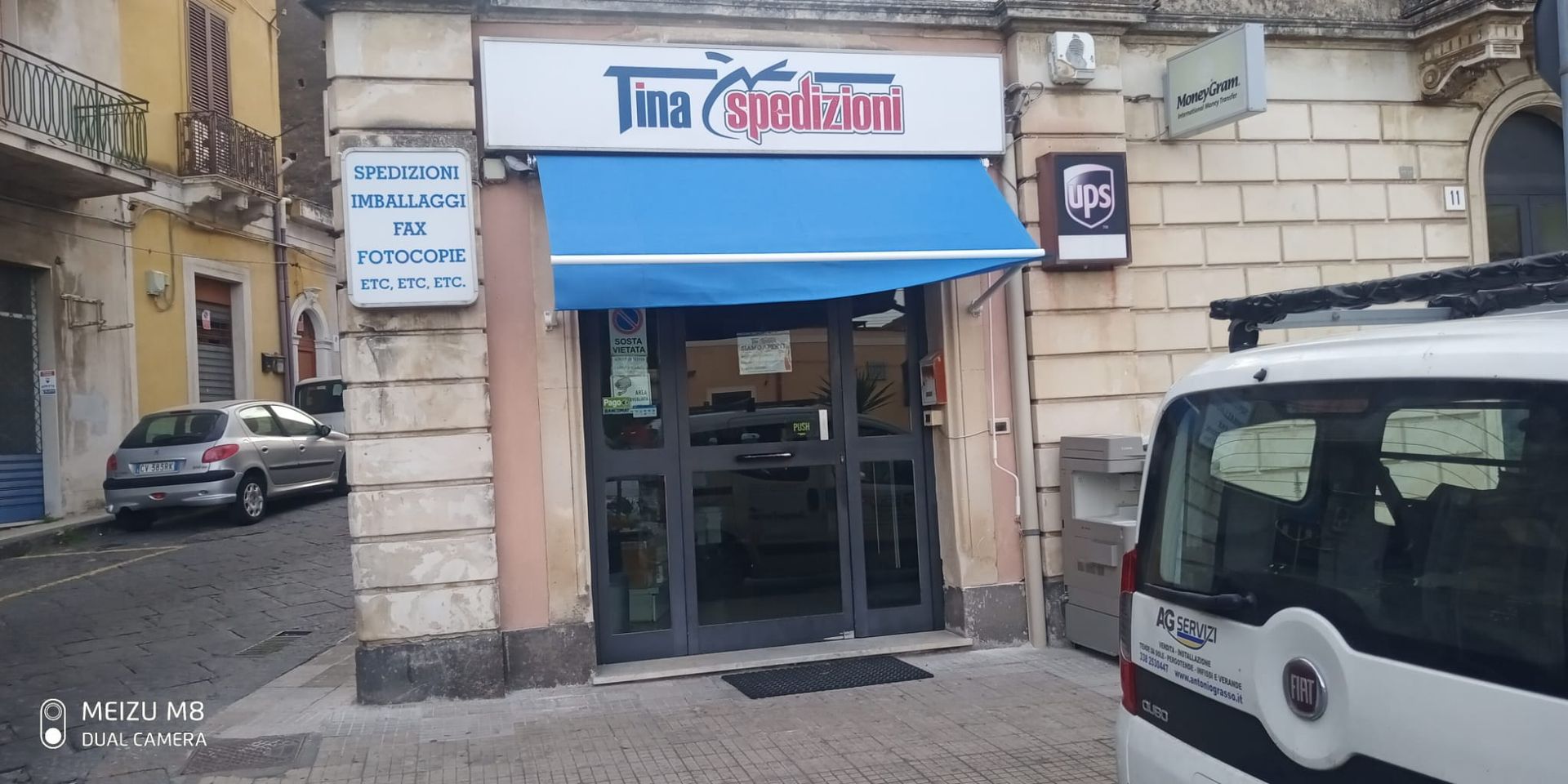Vetrina con tenda da sole blu e porta a vetri accanto a veicoli parcheggiati su una strada