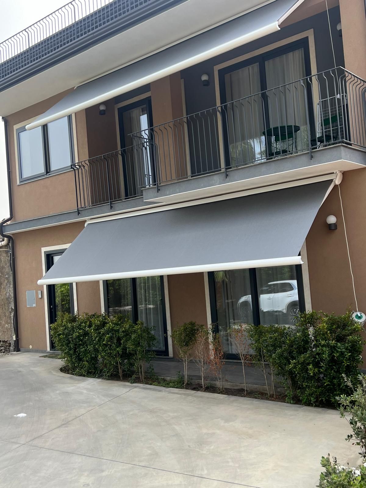Casa a schiera marrone a due piani con tende da sole grigie, balcone e arbusti lungo un vialetto in cemento.
