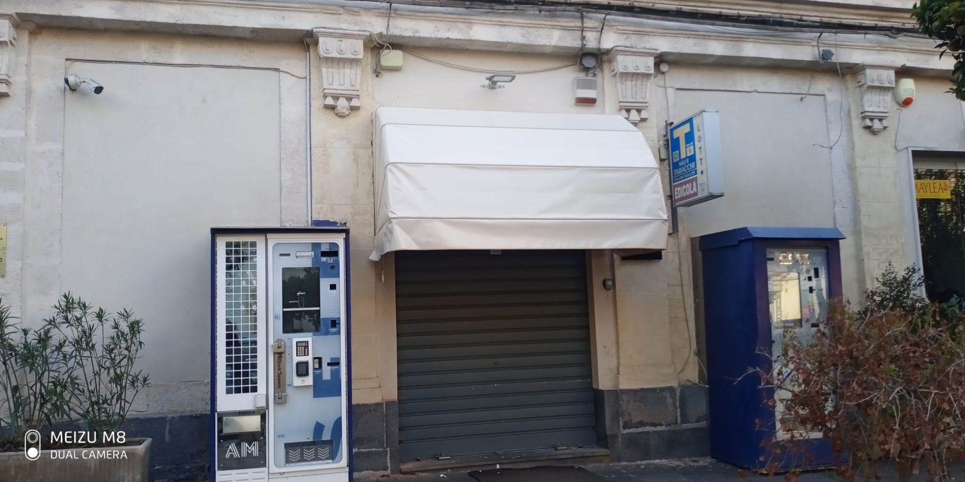 Negozio chiuso con serranda grigia, tenda da sole bianca e chiosco blu su una facciata stradale usurata