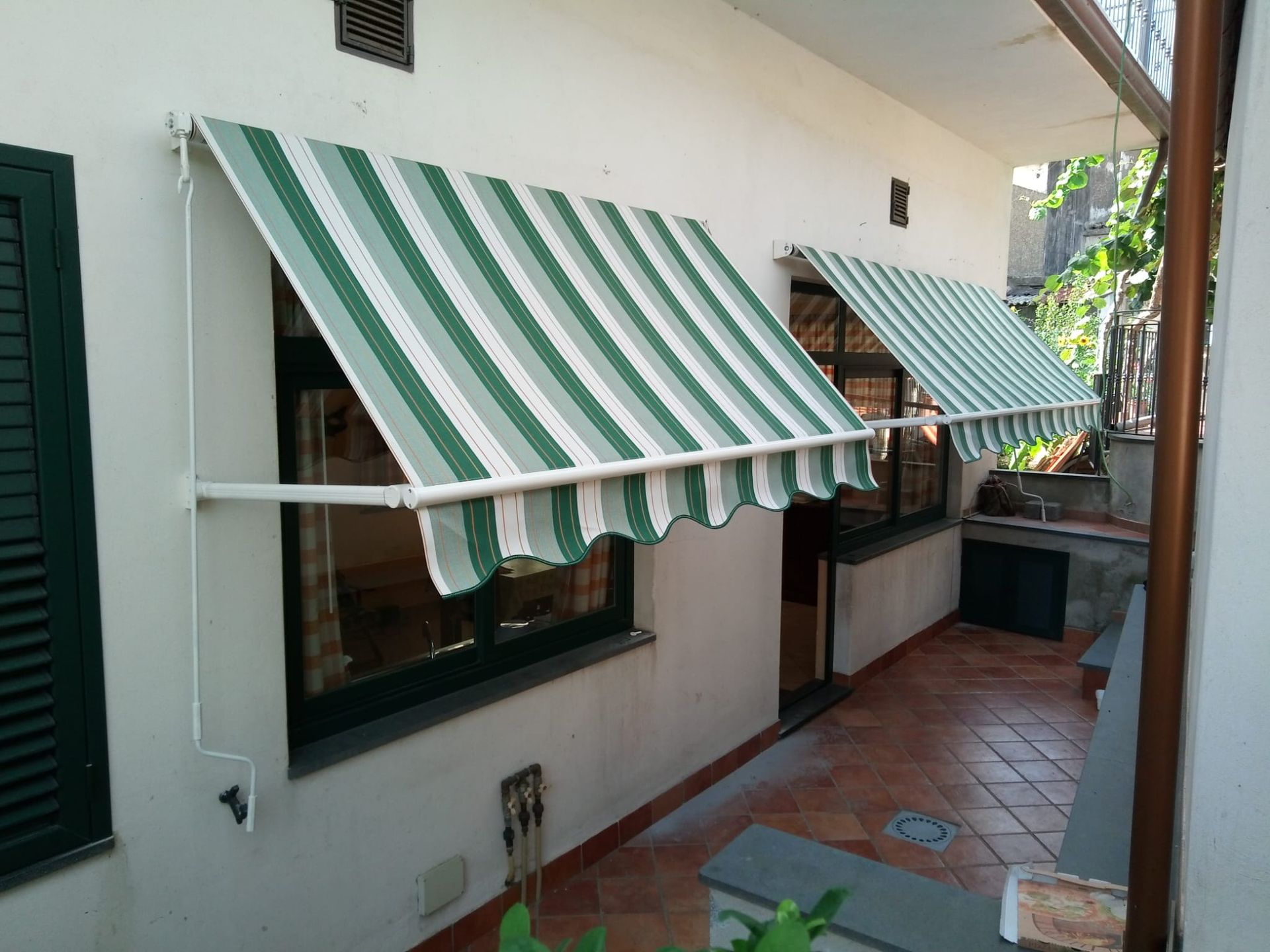 Esterno di una casa bianca con tende da sole a strisce verdi e bianche sopra le finestre e un vialetto piastrellato sul patio.