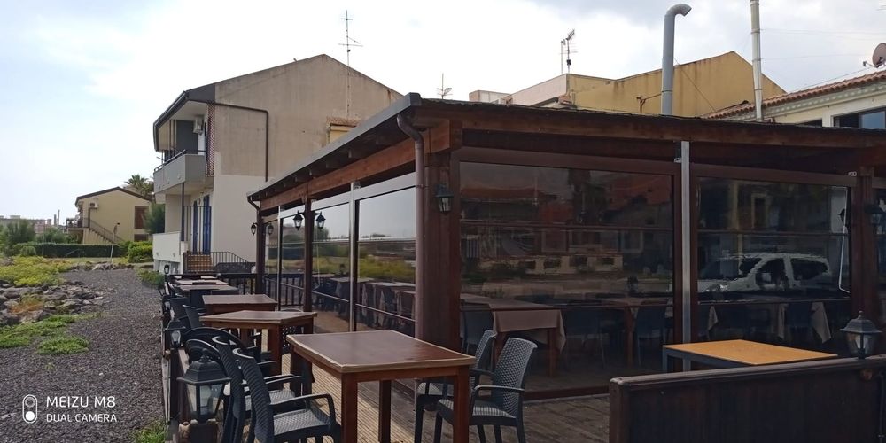 Tavolini e sedie all'aperto in legno sotto un patio coperto, accanto ad edifici e auto parcheggiate.