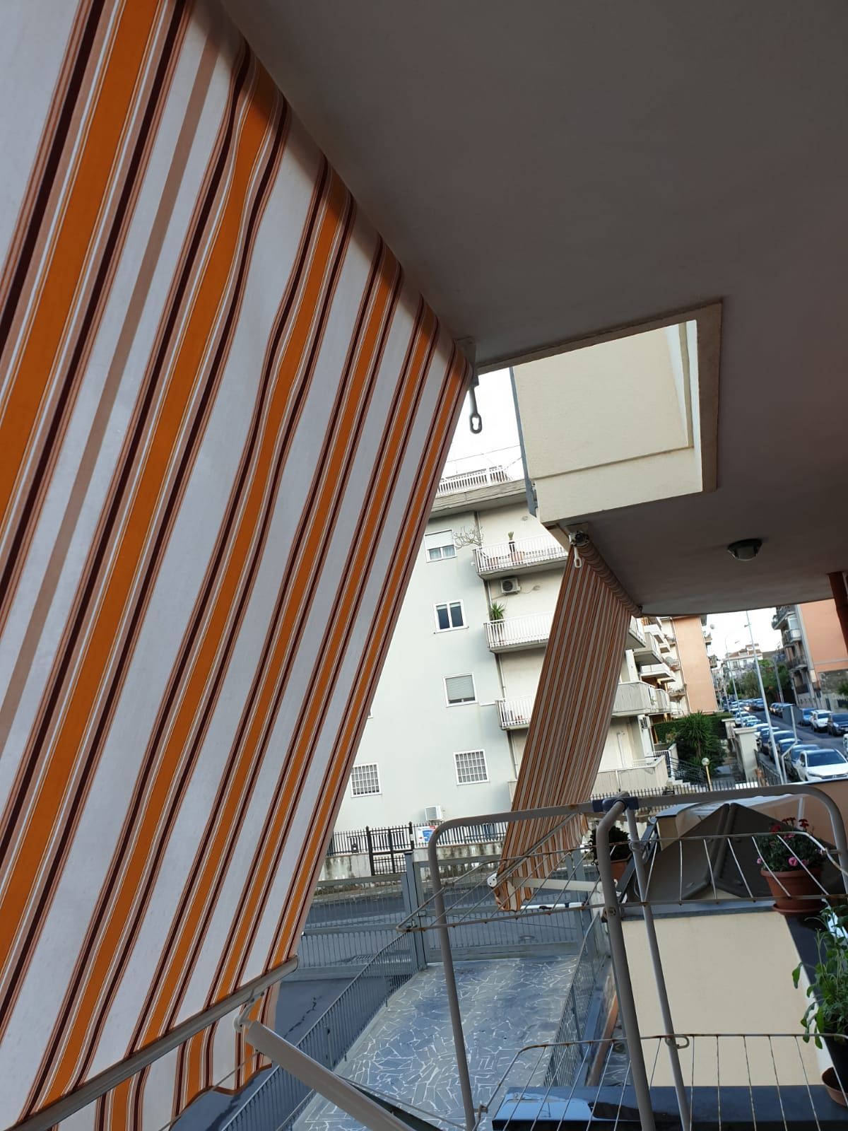 Balcone con tende a righe arancioni e bianche, affacciato sui palazzi circostanti e sulla strada.