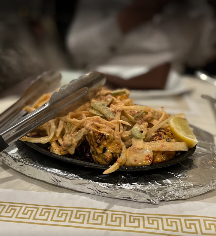 Sizzling Indian dish, possibly paneer tikka, on a platter with tongs and lemon slice.
