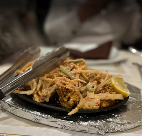 Sizzling plate of Indian food: grilled items, onions, peppers, creamy sauce, lemon wedge, tongs.