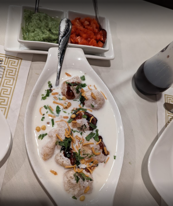 Dahi vada dish in white sauce, with side dishes of green and red sauces, on a white table.