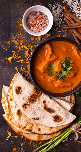 Chicken curry in black bowl, naan bread, spices, and rice on a dark surface.