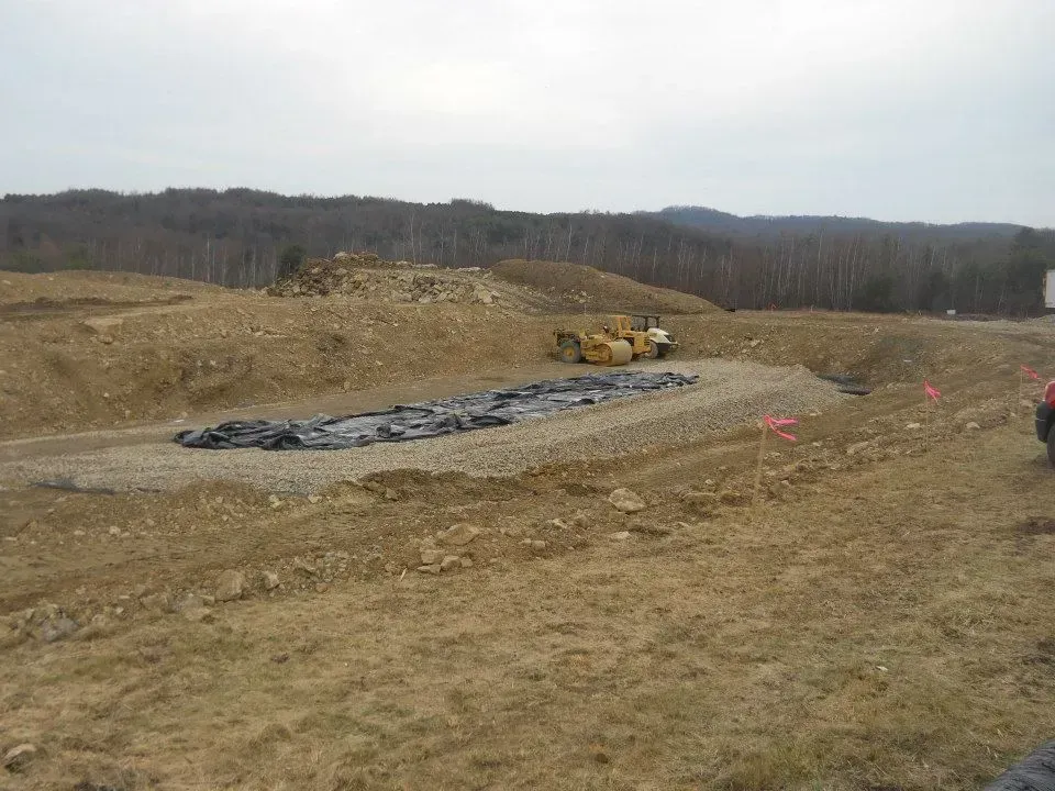Dirt construction site with excavator, tarp-covered trench, and hills in the background