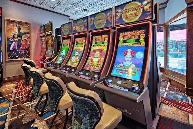 Row of brightly lit slot machines in a casino, with stools and daylight from windows.