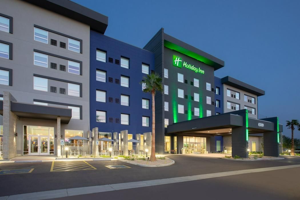 Exterior of a multi-story Holiday Inn hotel at twilight, featuring gray and blue panels with bright green branding.