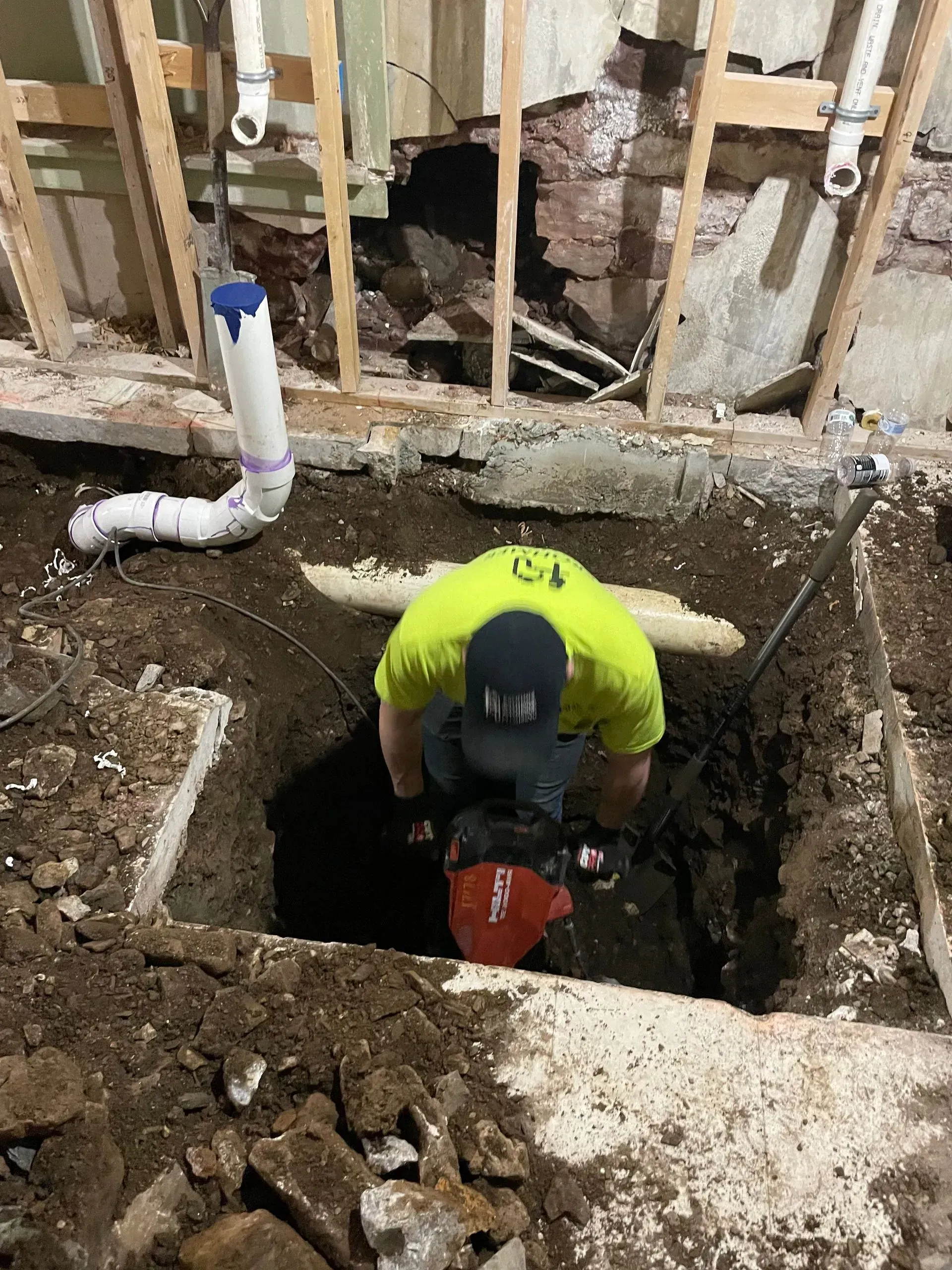 Worker in neon shirt crouches in a floor cutout during plumbing repair near exposed pipes and framing.