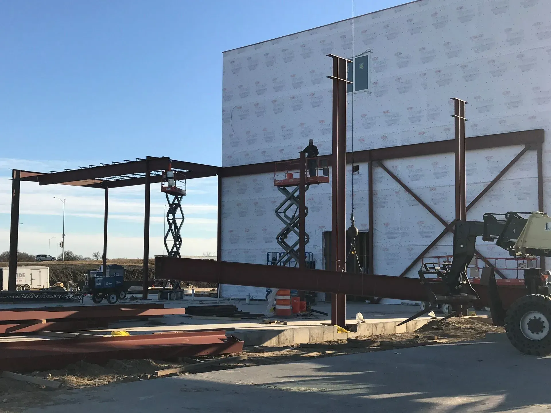 Steel frame structure being lifted by a forklift beside a white building on a construction site