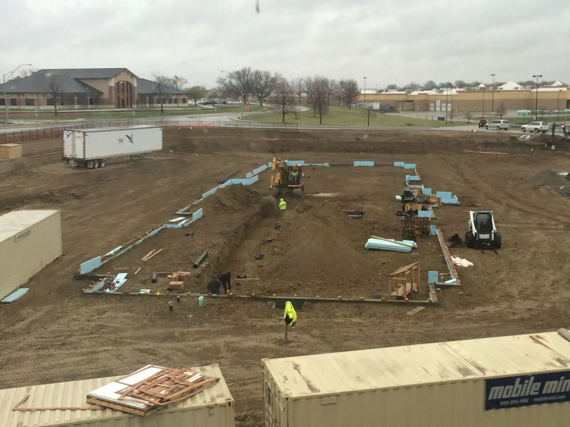 Construction site with excavated foundation, workers, and scattered blue insulation boards on muddy ground.