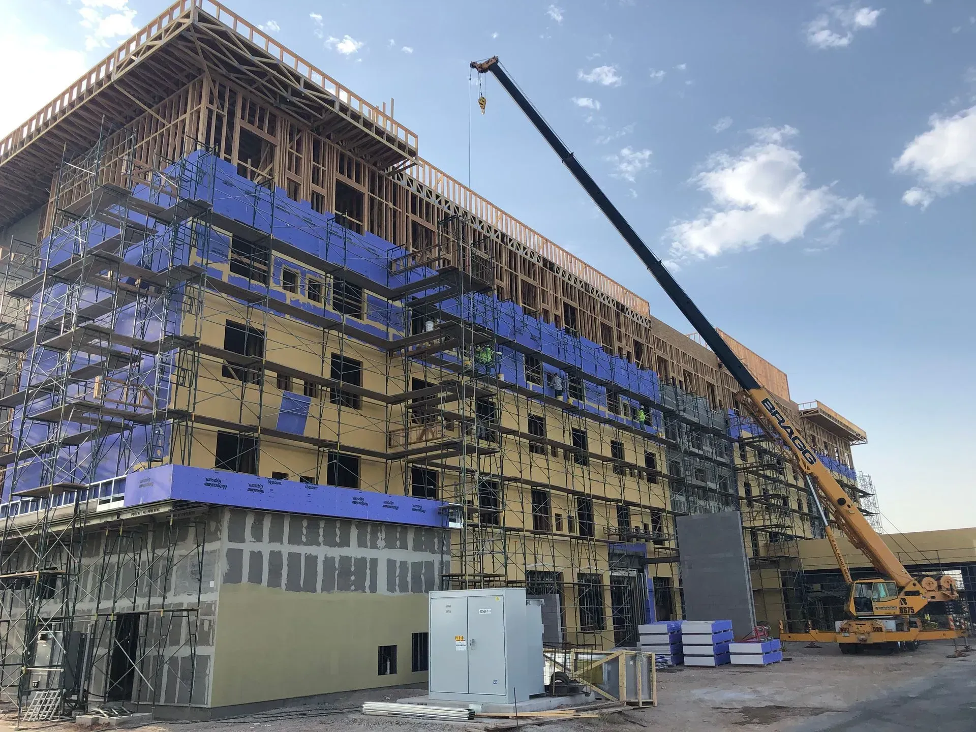 Multi-story building under construction with scaffolding, blue sheathing, and a crane on site