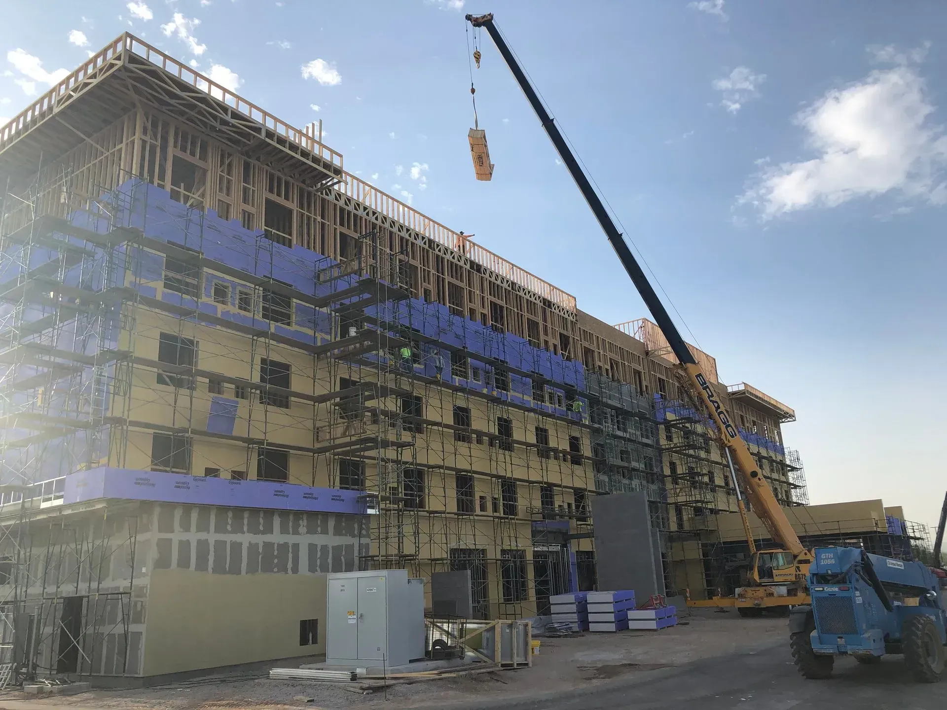 Construction site with multi-story building under scaffolding and a crane lifting materials nearby.