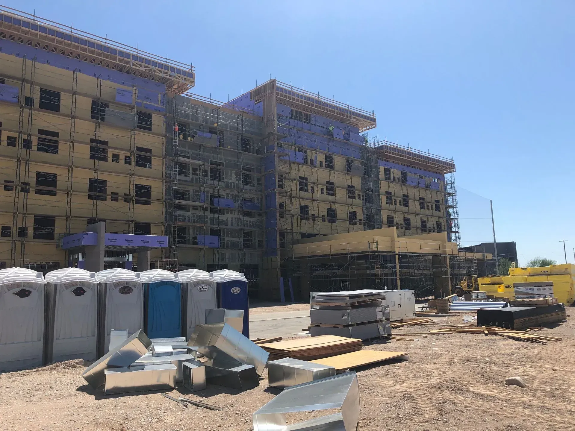 Construction site with a multi-story apartment building, portable toilets, and building materials under a clear blue sky